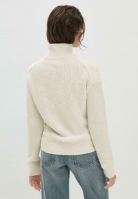 Frau mit welligem braunem Haar, die einen cremefarbenen, gestrickten Rollkragenpullover und eine blaue Jeans trägt, von hinten gegen einen schlichten Hintergrund gewandt.