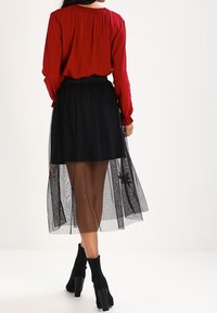 Femme portant un haut rouge, une jupe midi noire transparente brodée sur une mini-jupe, et des bottines à talons hauts noires, de dos.