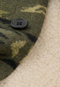 Green and black patterned wool fabric with a black four-hole button, resting on a textured beige surface.