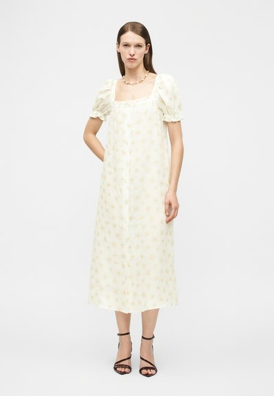 Γυναίκα στέκεται φορώντας λευκό μίντι φόρεμα με φουσκωτά μανίκια και κίτρινο λουλουδάτο μοτίβο, συνδυασμένο με μαύρα πέδιλα με λουράκια και ένα κολιέ.