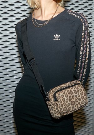 Femme portant une robe noire Adidas avec des rayures léopard sur les manches et un sac bandoulière léopard, devant un fond métallique géométrique.