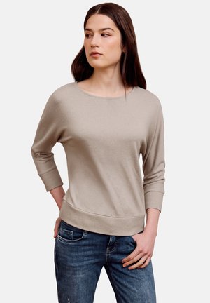 Junge Frau trägt einen beigen Langarmpullover und blaue Jeans, steht mit einer Hand in der Tasche und blickt leicht nach links.