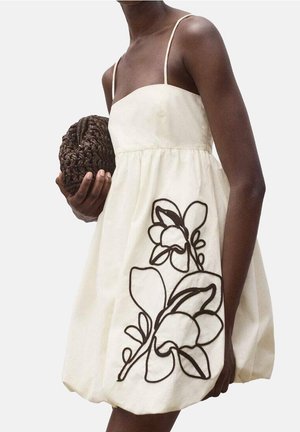 Frau in einem cremefarbenen Spaghettiträgerkleid mit auffälligem schwarzen Blumenmuster hält eine runde, geflochtene braune Clutch vor hellem, einfarbigem Hintergrund.