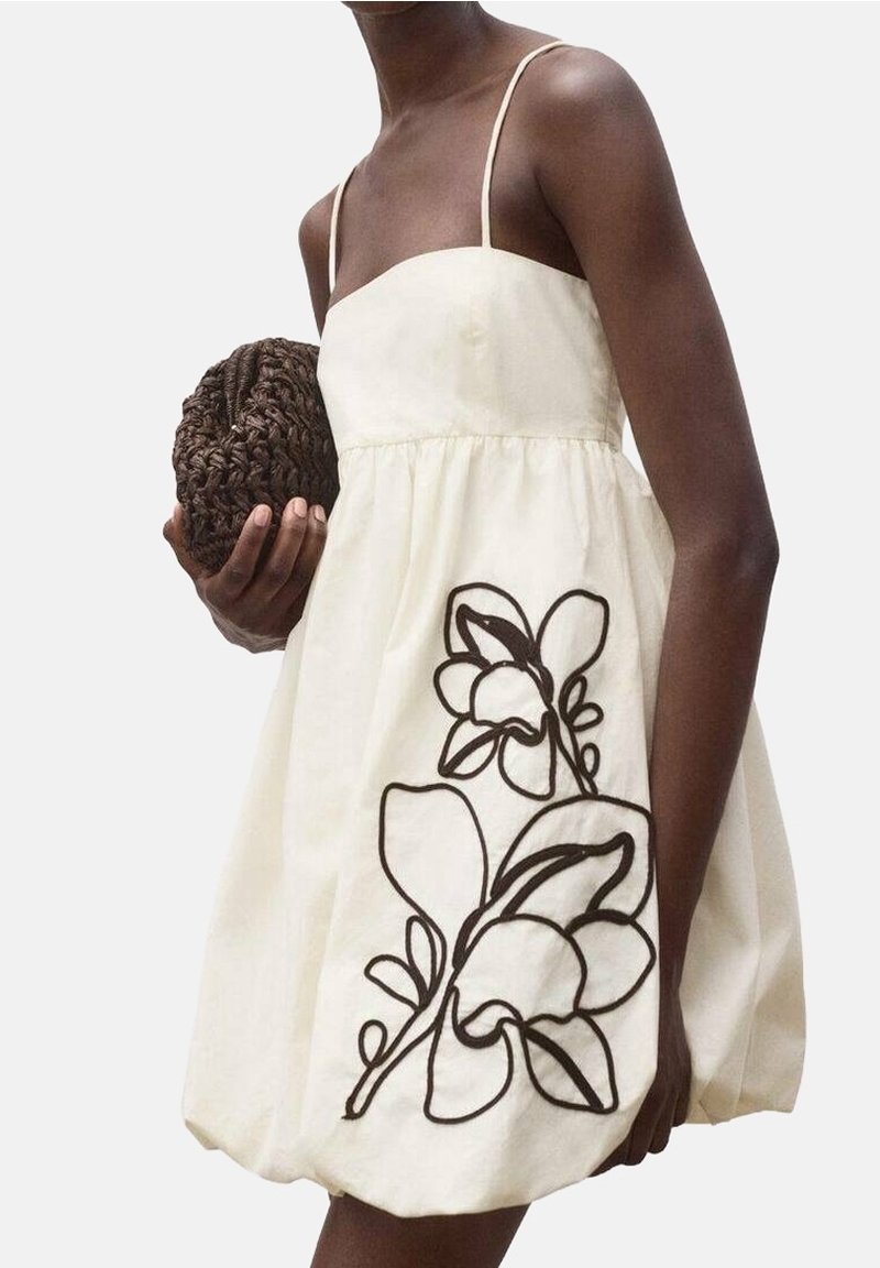 Femme portant une robe crème à fines bretelles avec un motif floral noir audacieux, tenant une pochette ronde tressée marron devant un fond uni clair.