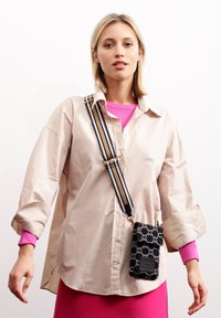 Beige übergroßes Hemd mit hochgerollten Ärmeln, darunter ein leuchtend pinkes Lagenhemd und eine schwarze gemusterte Handytasche mit einem gestreiften Schultergurt.