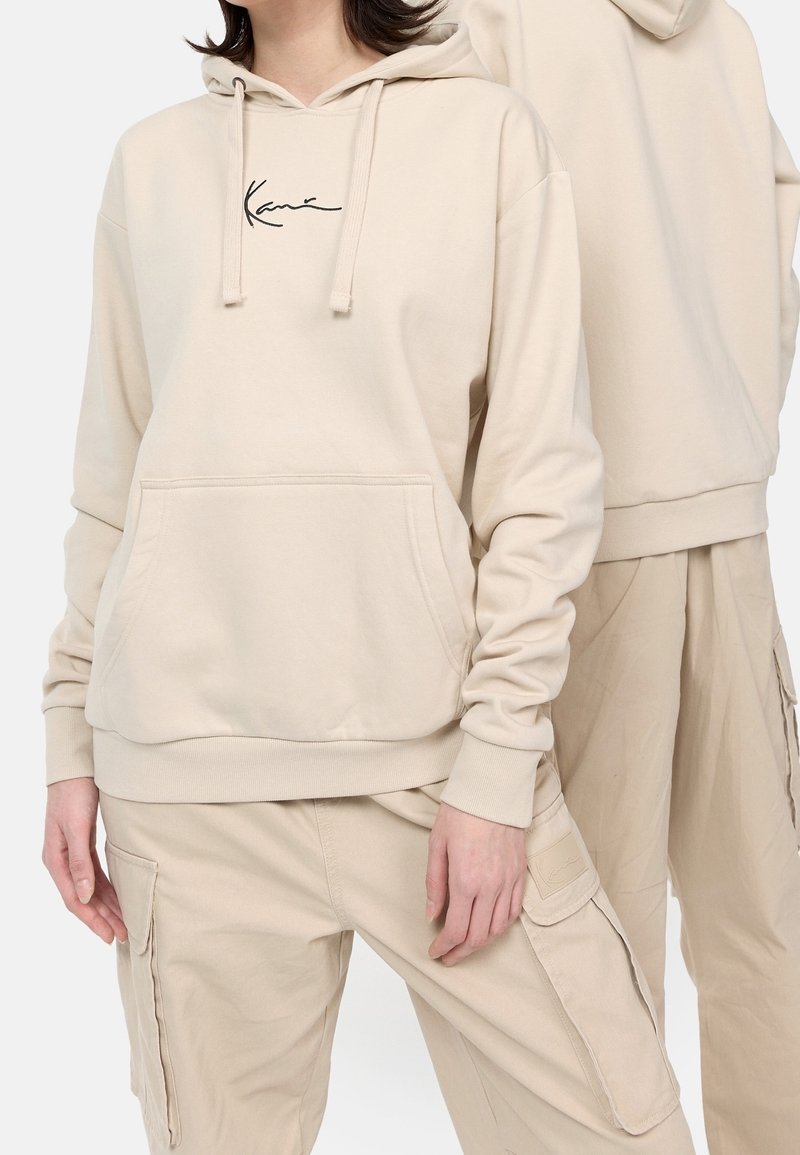 Felpa beige con cappuccio dotata di tasca a marsupio, logo ricamato nero sul petto, polsini e orlo a costine, abbinata a pantaloni cargo coordinati.