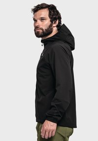 Schwarze wasserdichte Jacke mit Kapuze, mit Reißverschlusstasche, elastischen Bündchen und einem eleganten Design, getragen über grünen Hosen.