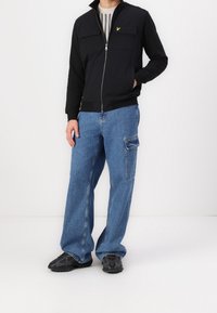 Chaqueta negra con cremallera y dos bolsillos en el pecho, combinada con pantalones cargo de mezclilla azul de corte holgado que tienen un bolsillo lateral. Zapatillas deportivas negras.