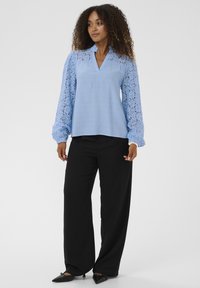 Haut bleu clair en dentelle, manches longues bouffantes et col en V ; associé à un pantalon noir à jambes larges et des ballerines noires pointues. Texture lisse.