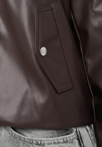 Brūna ādas jaka ar atloku kabatu un sudraba metāla detaļām, kombinēta ar gaiši pelēcīgiem džinsiem kontrastam. Teksturēta ādas virsma.