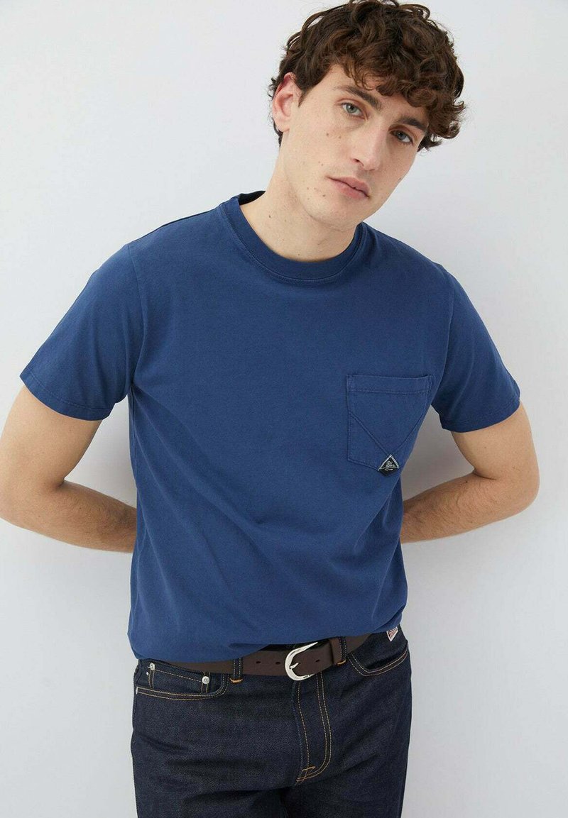 T-shirt blu navy a maniche corte con taschino sul petto, colletto tondo, realizzato in cotone, abbinato a jeans scuri e una cintura marrone.