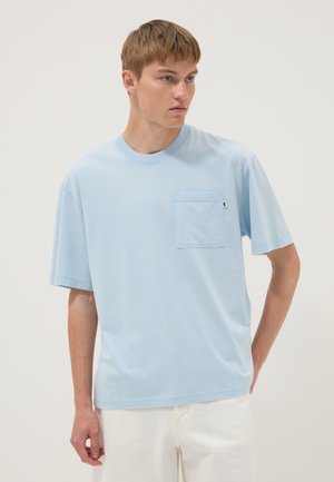 Joven con camisa azul claro de manga corta con bolsillo en el pecho, mirando hacia un lado sobre un fondo liso.