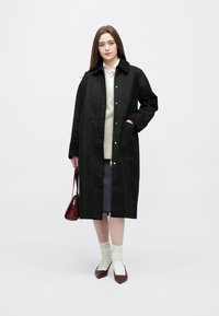 Palton lung negru, din material rezistent, cu închidere frontală cu nasturi, două buzunare laterale și guler clasic, purtat peste un pulover deschis la culoare.