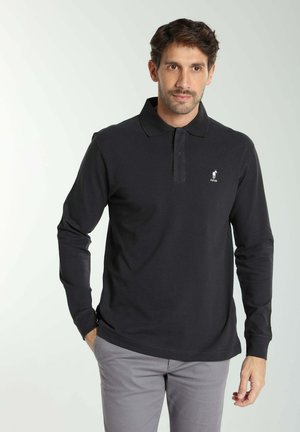 Homme portant un polo noir à manches longues avec un petit logo blanc et un pantalon gris clair, debout avec une main dans la poche sur un fond clair.