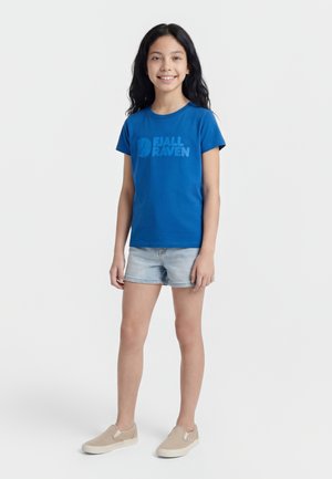 Young girl with long black hair smiling, wearing a blue Fjällräven t-shirt, light denim shorts, and beige slip-on shoes, standing on white background.