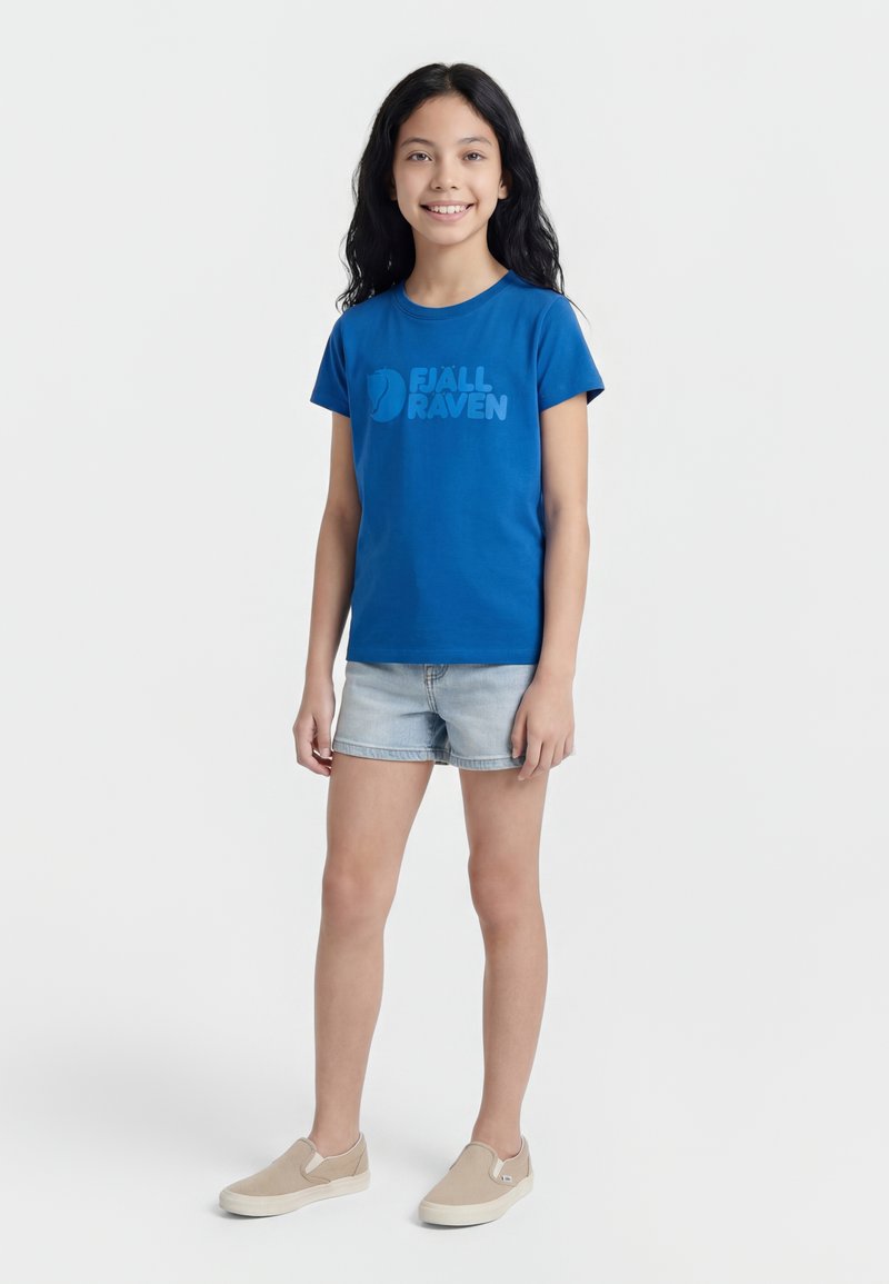 Jeune fille aux longs cheveux noirs souriante, portant un t-shirt bleu Fjällräven, un short en jean clair et des chaussures beige à enfiler, debout sur fond blanc.
