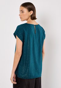 Nainen seisoo sivuttain, päällään teal-värinen haalariblouse, jossa on zikkurat kuvio ja avonainen selkä, sekä löysät mustat housut valkoista seinää vasten.
