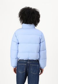 Svetlomodrá bunda typu puffer s vysokým golierom, prešívaným dizajnom a elastickými manžetami. Kombinovaná s tmavými džínsami, zobrazená zozadu.