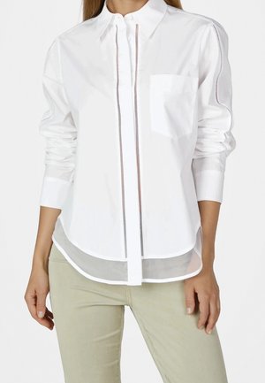 Femme portant une chemise blanche à manches longues avec une poche poitrine devant et un pantalon beige, debout, les mains détendues le long du corps.