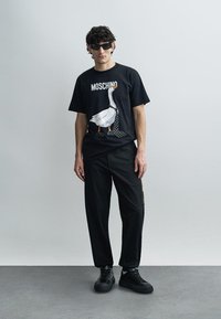 Fekete Moschino póló libamintával és fehér felirattal, laza fekete nadrággal és fekete sportcipővel párosítva. Sima anyag és casual szabás.