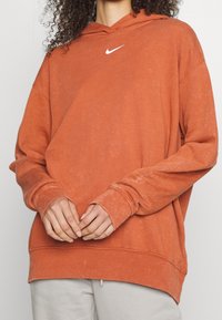 Oranžová mikina s kapucí se bílým logem Nike na hrudi. Má dlouhé rukávy, volný střih a mírně asymetrický lem.