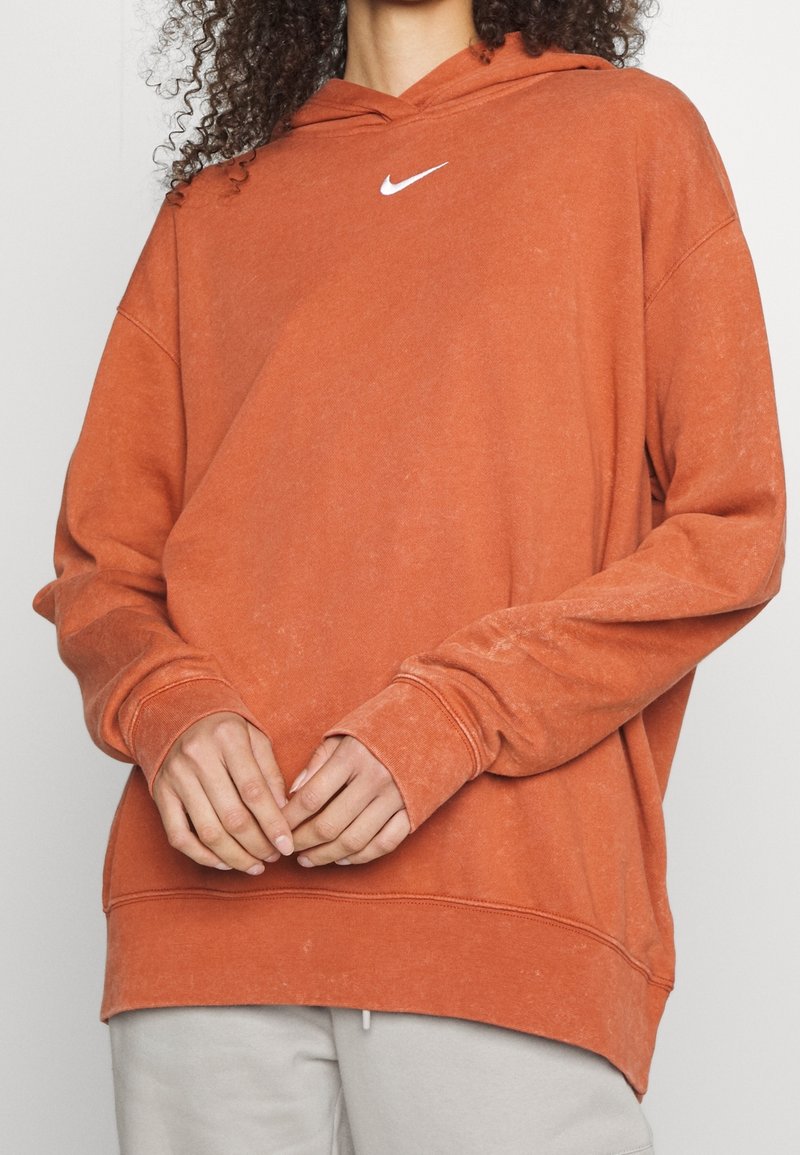 Oranžová mikina s kapucí se bílým logem Nike na hrudi. Má dlouhé rukávy, volný střih a mírně asymetrický lem.