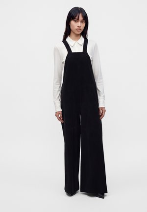 Jeune femme aux longs cheveux foncés portant une chemise blanche à manches longues avec col et une salopette noire à jambes larges, debout devant un fond uni.