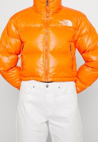 Jasnopomarańczowa kurtka puchowa z wysokim kołnierzem, zamkiem błyskawicznym i logo na lewej piersi, w połączeniu z białymi spodniami. Gładka, błyszcząca tekstura.