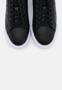 Paire de baskets en cuir noir avec lacets noirs et semelles en caoutchouc blanc, vues de dessus sur un fond gris clair.