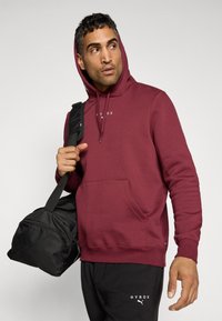 Bordeaux hættetrøje med lommer foran, snor i hætten og hvidt logo-tekst. Parret med sorte sweatpants og holder en sort duffelbag.