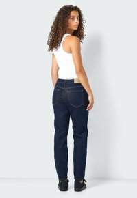 Temno prani jeans hlače s ravnim krojem, ki imajo pet žepov in kontrastno šivanje. Ujemajo se z belo topom brez rokavov in temnimi čevlji.
