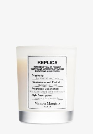 Bougie parfumée étiquetée "Replica" par Maison Margiela, parfum "Au coin du feu", notes de bois brûlé et de châtaigne, support en verre blanc.