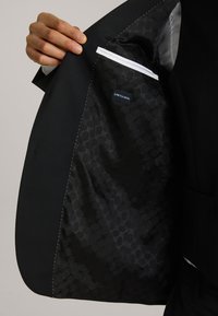 Das Innere der schwarzen Sakkojacke hat ein glattes, gemustertes Futter mit dem Etikett "STRETCH WOOL". Mit weißen Steppnähten akzentuiert.
