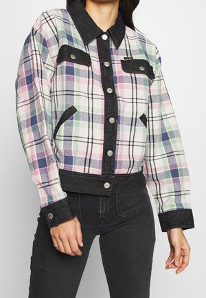 Mujer con chaqueta de botones con estampado de cuadros en tonos pastel, con cuello negro, puños negros y ribete negro, combinada con vaqueros negros.