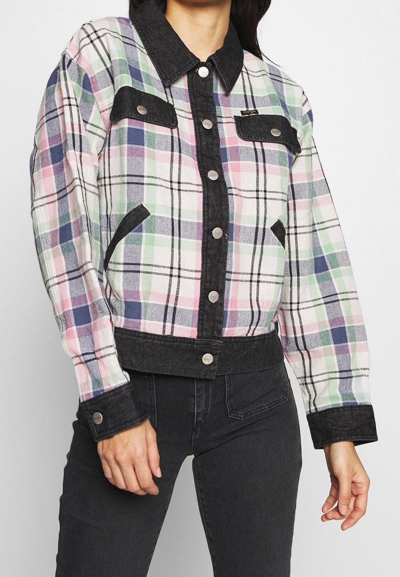 Femme portant une veste à carreaux pastel boutonnée avec col noir, poignets noirs et bordure noire, associée à un jean noir.
