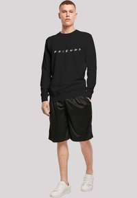 Svart hettegenser med "FRIENDS"-logo i hvitt, kombinert med sorte shorts. Modellen har på seg hvite sneakers og demonstrerer en avslappet atletisk stil.