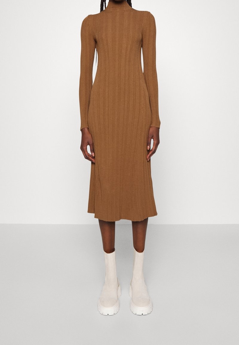 Femme portant une robe midi marron côtelée à manches longues avec un col montant et des bottines blanches épaisses, debout devant un fond uni.