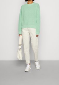 Pullover a maglia verde menta, pantaloni bianchi e sneakers bianche. Porta una piccola borsa bianca. Outfit casual con texture e linee pulite.