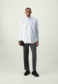 Homem vestido com uma camisa branca de botões, calças cinzas, tênis pretos, segurando uma bolsa clutch preta, posando contra um fundo liso.