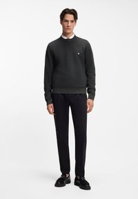 Dunkelgrauer, strukturierter Pullover mit Rundhalsausschnitt, getragen über einem weißen Hemd mit Kragen. Kombiniert mit schwarzen, maßgeschneiderten Hosen und schwarzen Loafers.