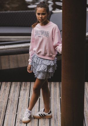 Lichtroze sweatshirt met de tekst "HOLLYWOOD", gelaagde blauwe gemusterde rok en zwart-witte sneakers, staande op houten planken.