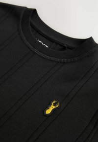 Sort sweatshirt med ribbet krave, der har vertikale sømdetaljer og et gult broderet hummerpatch på venstre bryst.