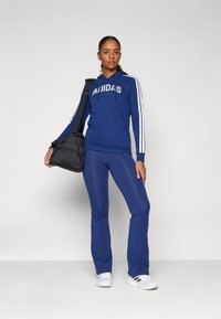 Felpa con cappuccio blu con logo e strisce bianche, abbinata a leggings blu e scarpe da ginnastica bianche. Borsa nera appesa alla spalla.