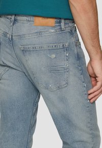 Jasnoniebieskie dżinsy wyróżniają się prostym krojem nogawki, subtelnymi przetarciami oraz skórzaną metką marki na pasku.