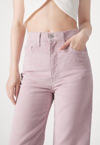 Pantalones de pana color rosa claro con talle alto, que presentan un bolsillo delantero, presillas para cinturón y un detalle de botón plateado. Textura suave, corte de pierna recta.