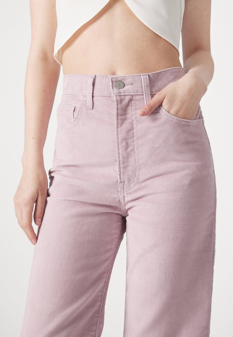 Pantalones de pana color rosa claro con talle alto, que presentan un bolsillo delantero, presillas para cinturón y un detalle de botón plateado. Textura suave, corte de pierna recta.
