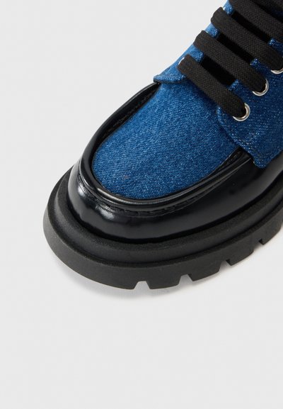 Bota de tobillo de denim azul con detalles en cuero negro brillante, suela de goma gruesa y cordones negros; presenta una puntera redondeada y tejido texturizado.