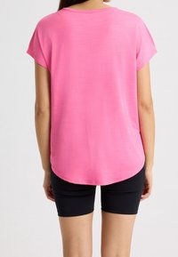 Top de manga corta y cuello redondo en un tono rosa brillante, fabricado en una tela ligera. Presenta un dobladillo curvado y un corte relajado, combinado con pantalones cortos negros.