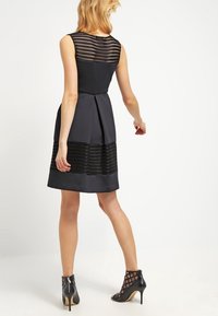 Robe noire avec un empiècement en crochet texturé et une jupe plissée. Fini avec des accents noirs transparents et accompagné de chaussures à talons hauts noires.