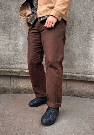 Brown cotton pants with side pockets, paired with black textured athletic shoes. The outfit features a tan outer jacket and patterned shirt.